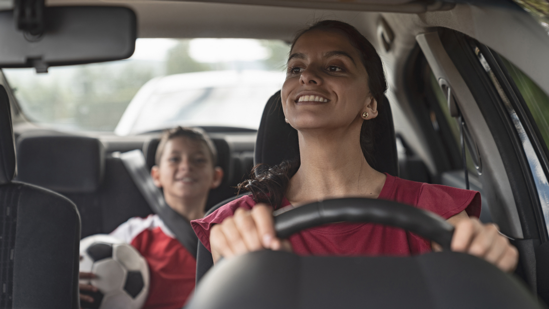 Child with soccer ball sits in the back seat of a car. Mother is in the driver's seat looking back at him